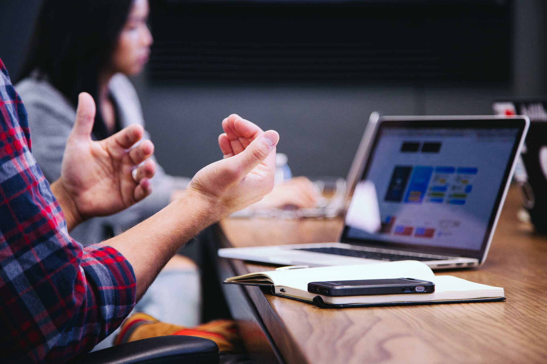 A close-up of gesturing hands belong to a man who is speaking in a meeting with a laptop in front of him and a woman in the background.