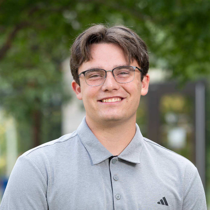 A smiling man with short brown hair and glasses wears a light gray Adidas polo against a blurred outdoor background