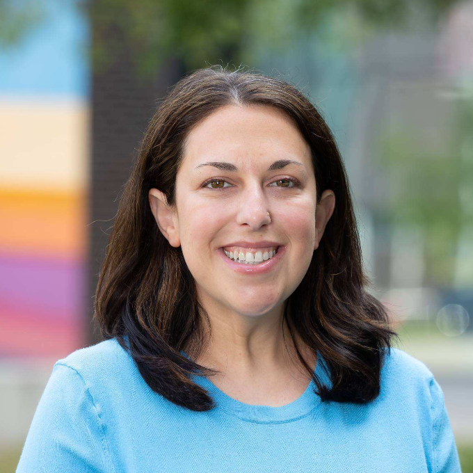 A smiling woman with shoulder-length dark brown hair wears a light blue sweater against a softly blurred outdoor background.