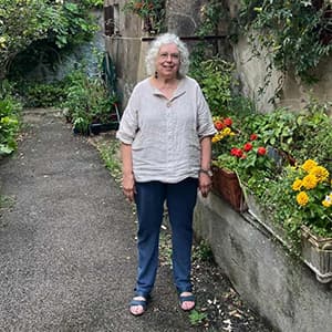 Woman with shoulder-length white hair wearing a light-colored blouse and jeans smiles while standing in front of a garden.