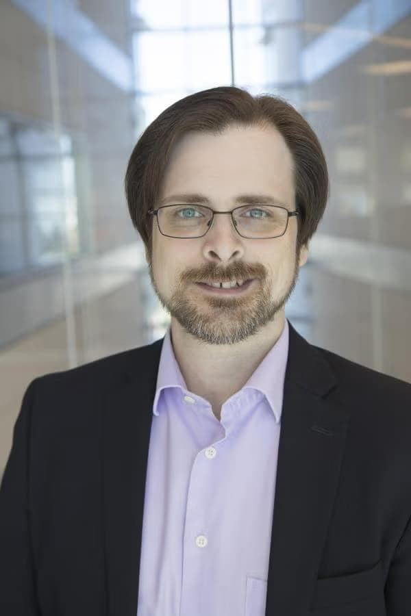Dr. Vose in a button-up and jacket and wire-framed glasses with a one-sided smile showing his teeth.