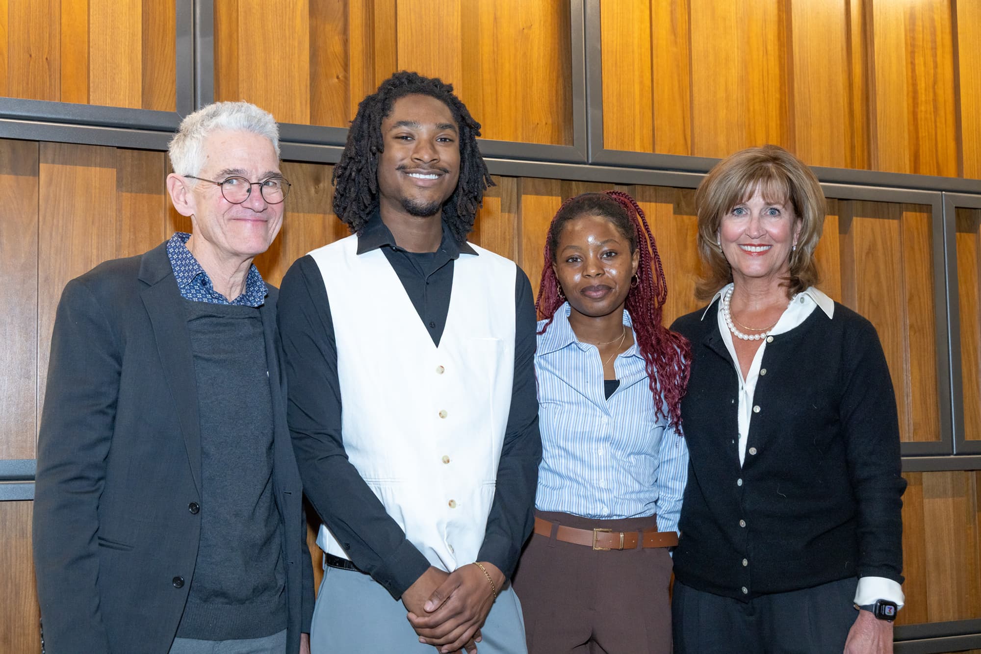 Four people in business professional clothing smile in front of a wood wall.