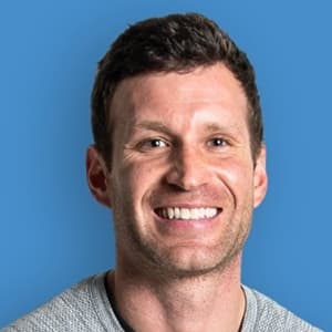 Headshot of a man in a gray shirt and smiling with his teeth against a bright blue background.