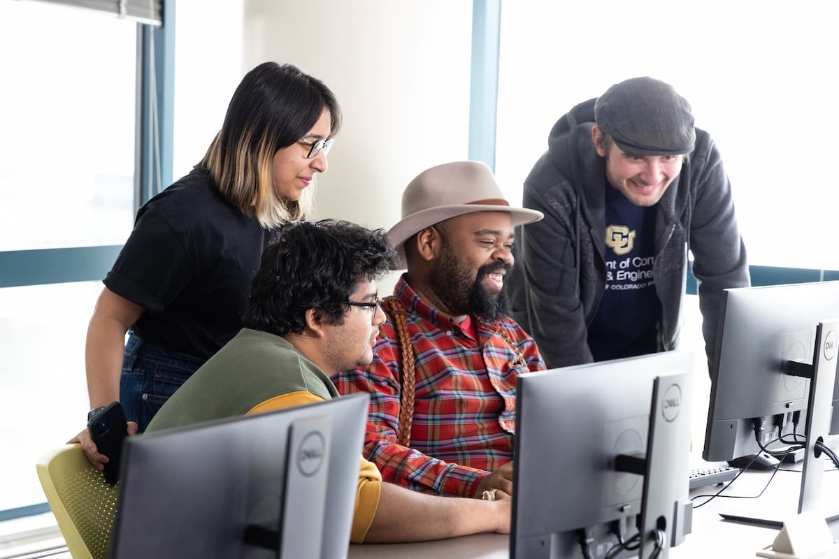 Group of four people, two seated, two standing, engaging with each other as they look at a computer screen.