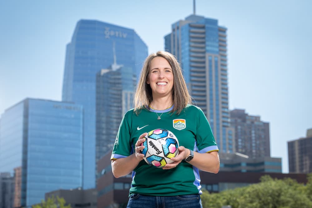 Woman smiling with shoulder-length hair wearing a green Denver Summit FC jersey and holding a soccer ball.