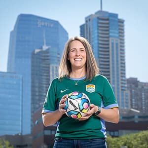 Woman smiling with her teeth wearing a green Denver Summit FC shirt and holding a soccer ball.