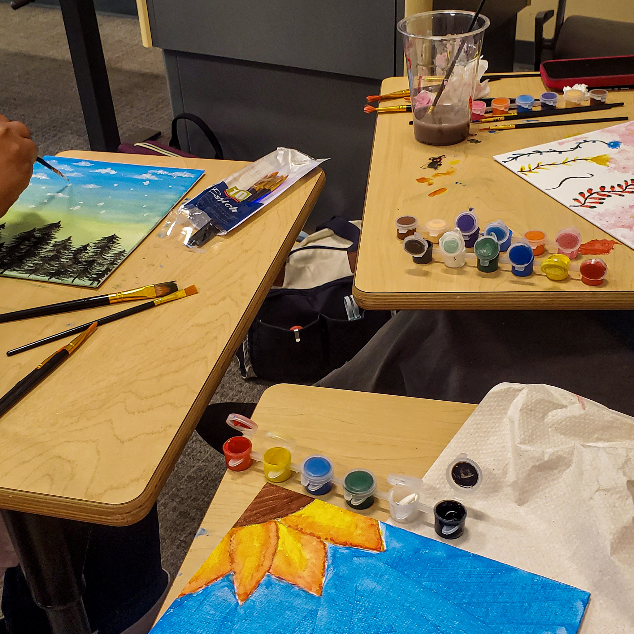 Students participate in a painting event (Sip & Paint), with colorful artwork, paintbrushes, and small pots of paint spread across tables.
