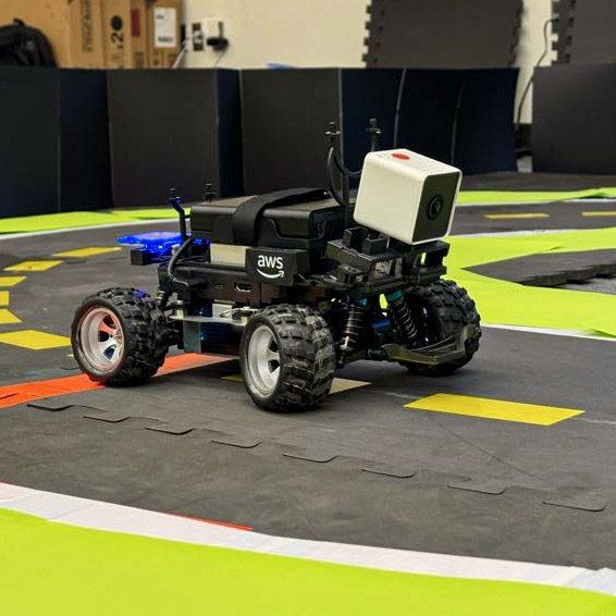 An AWS DeepRacer car sits on the track during the CEDC AI Grand Prix 2026 at CU Denver. The small autonomous vehicle is equipped with a camera and positioned on a course marked with bright yellow and green lines.