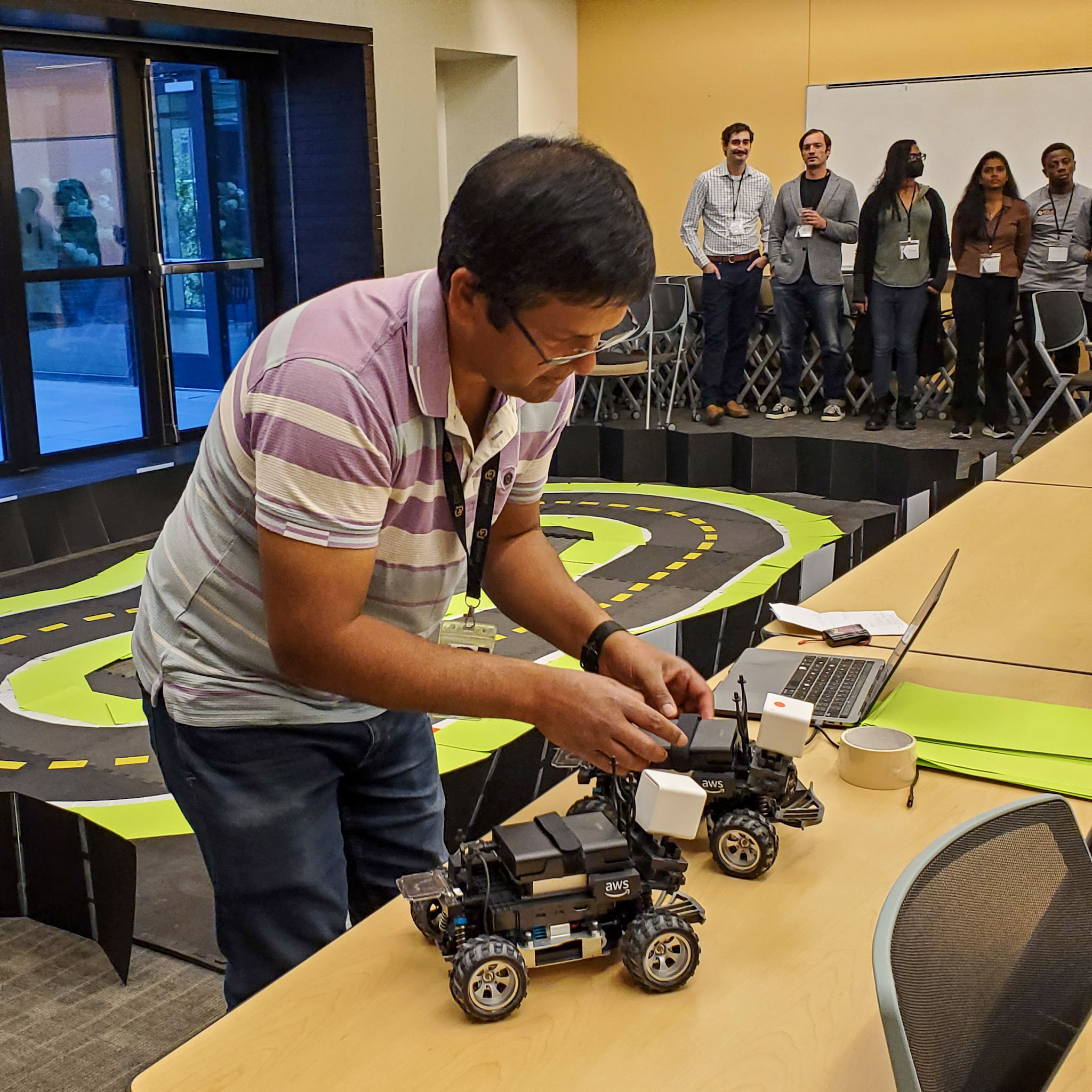 Dr. B, organizer of the CEDC AI Grand Prix 2026 student competition at CU Denver, adjusts an AWS DeepRacer car beside a second car on a table. A track for testing autonomous driving models is set up nearby as event participants watch.