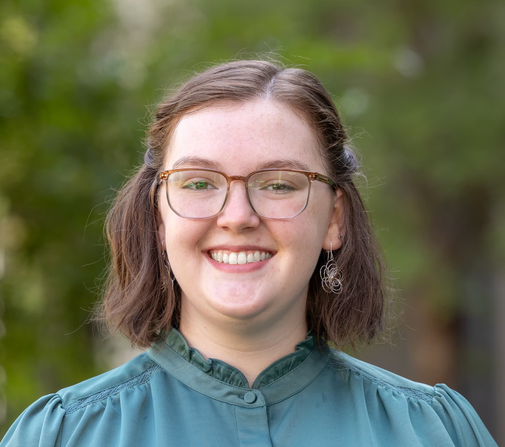 Emily, a caucasian woman with brown hair and glasses, stands in front of a tree.