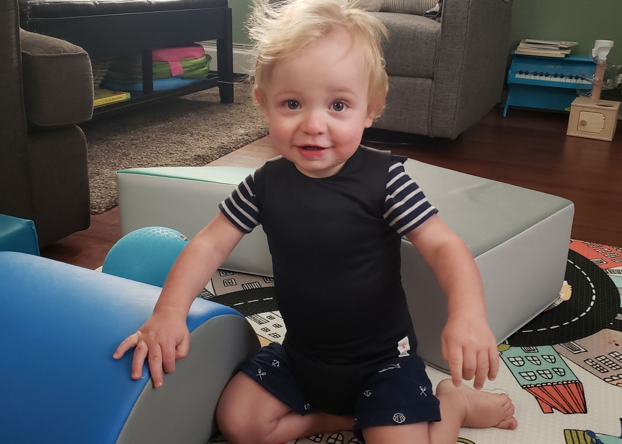 A toddler smiles kneeling with his toys wearing a black vest.